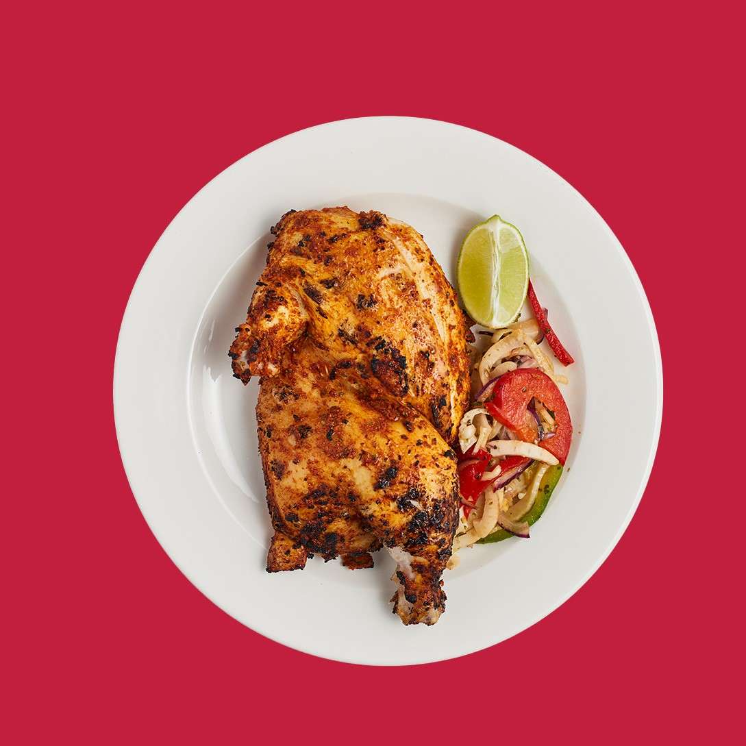 Roasted half chicken rests on a white plate, accompanied by sautéed onions and red and green peppers plus a lime wedge, set against a solid red background.