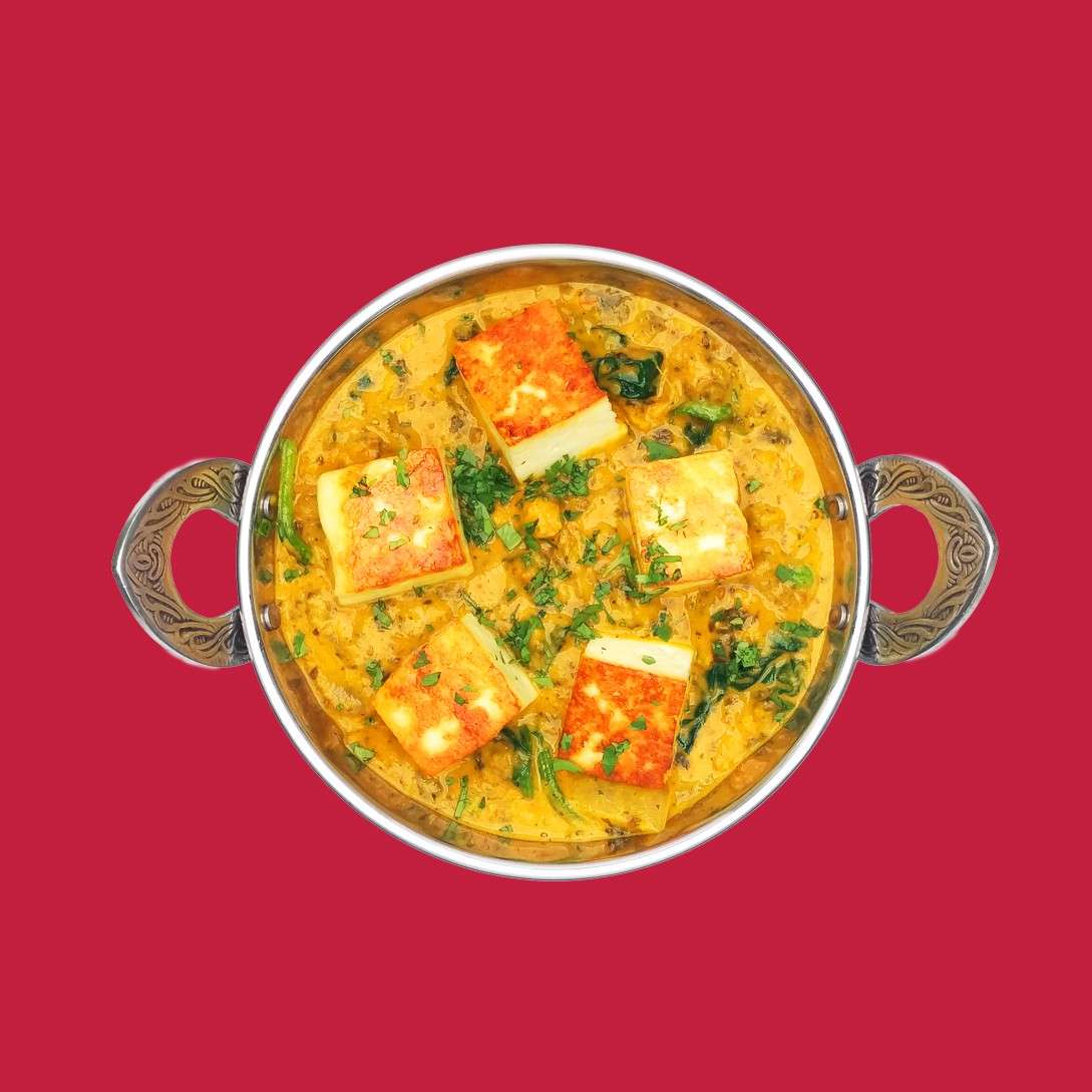 A metal-handled bowl presents paneer cubes simmering in creamy yellow curry with spinach and herbs, viewed from above, set against a plain deep-red background.