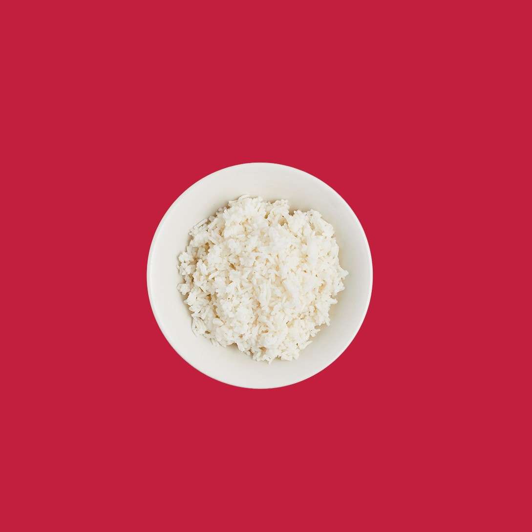 A white bowl of cooked rice sits centered, viewed from above, against a solid red background with ample empty space.
