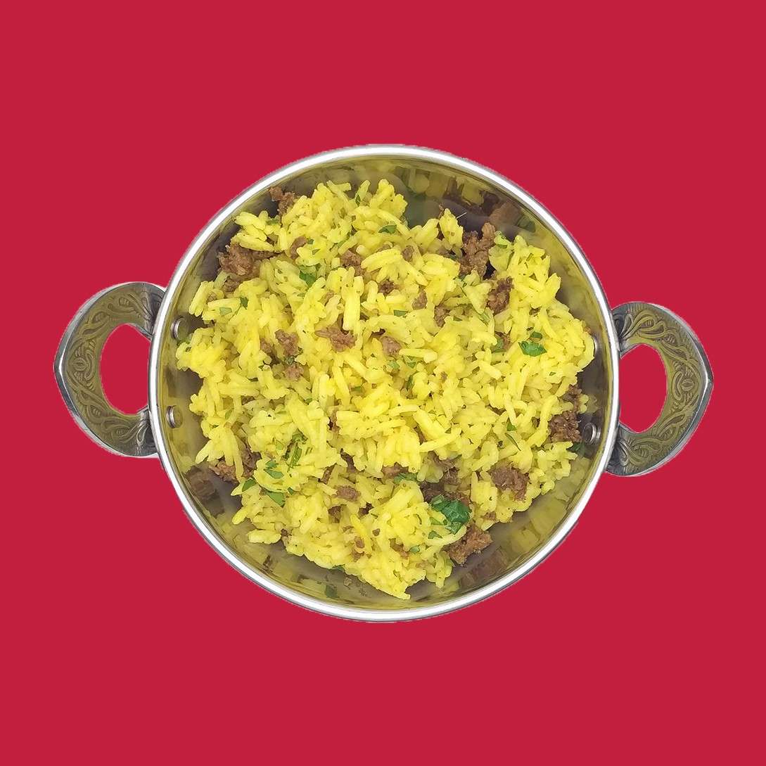 Bowl of yellow rice rests, garnished with herbs and mixed with small meat pieces, in a silver handled pot, centered against a plain red background.