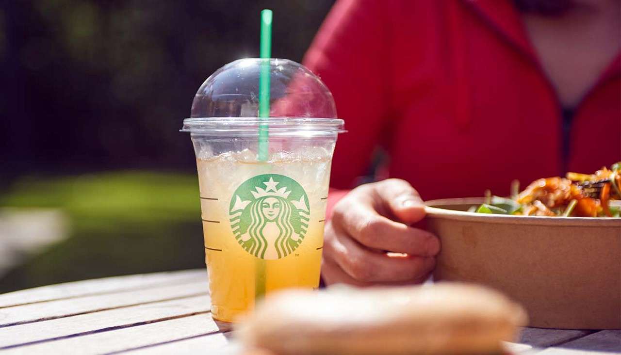 Plastic cup of iced drink with green straw rests on an outdoor table, condensation beading; nearby, a person in a red top eats from a cardboard bowl, greenery blurred behind.
