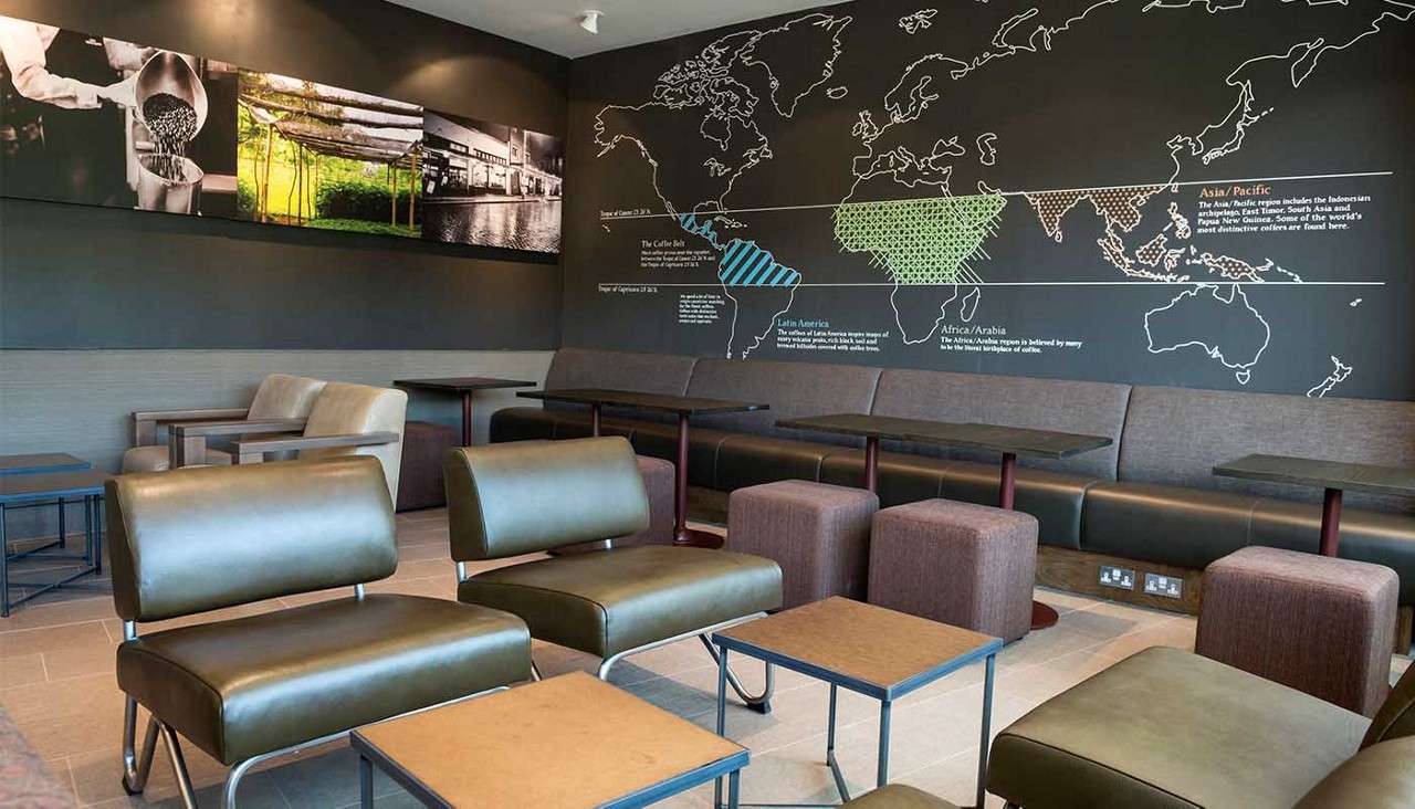 Lounge chairs sit arranged around low tables; a long banquette lines the wall beneath a world map highlighting coffee regions in a modern café.
Text: Latin America; Africa/Arabia; Asia Pacific; The Coffee Belt; Tropic of Cancer; Tropic of Capricorn; additional small descriptive text unreadable.