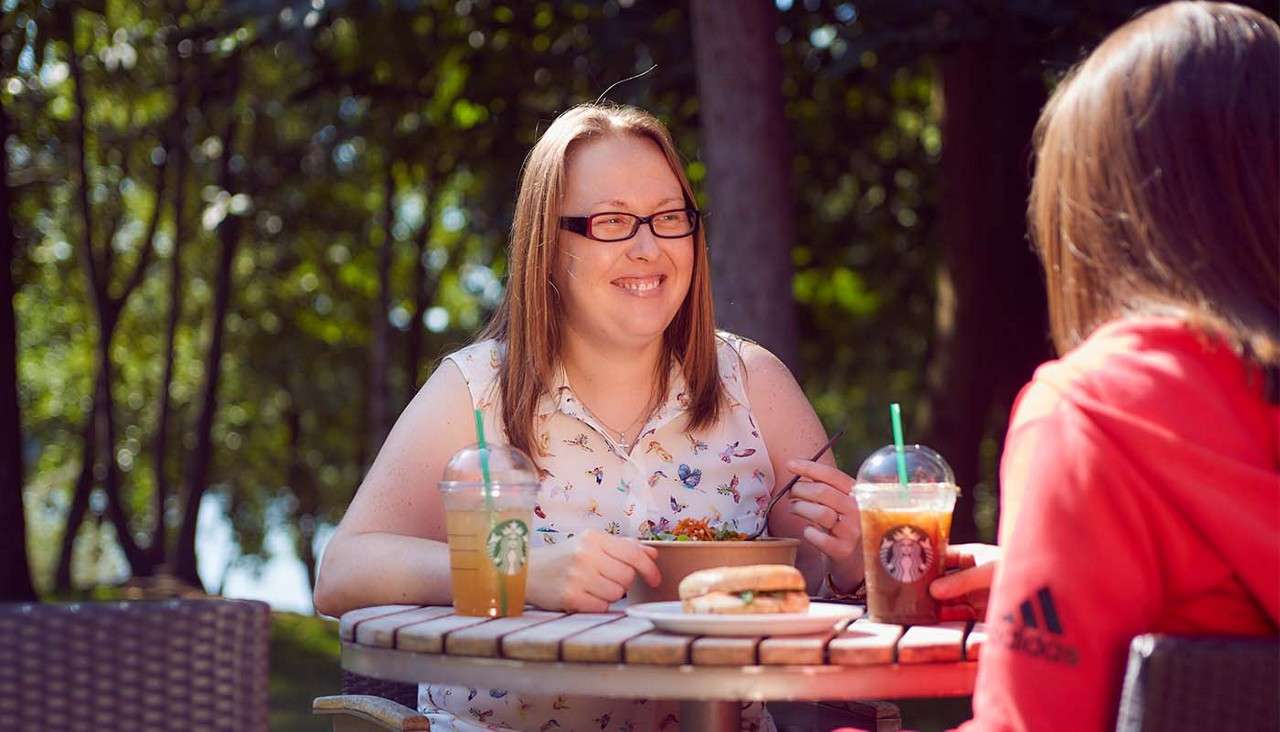 Two people chat while eating at a round outdoor café table; one smiles over a salad. Iced drinks and a sandwich sit nearby. Text: STARBUCKS; adidas.
