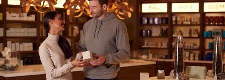 Two shoppers hold and discuss a box, smiling. Warm pendant lights glow as shelves display skincare in a boutique. Backlit signage reads: ELEMIS. Glass cylinders and jars decorate the counter.