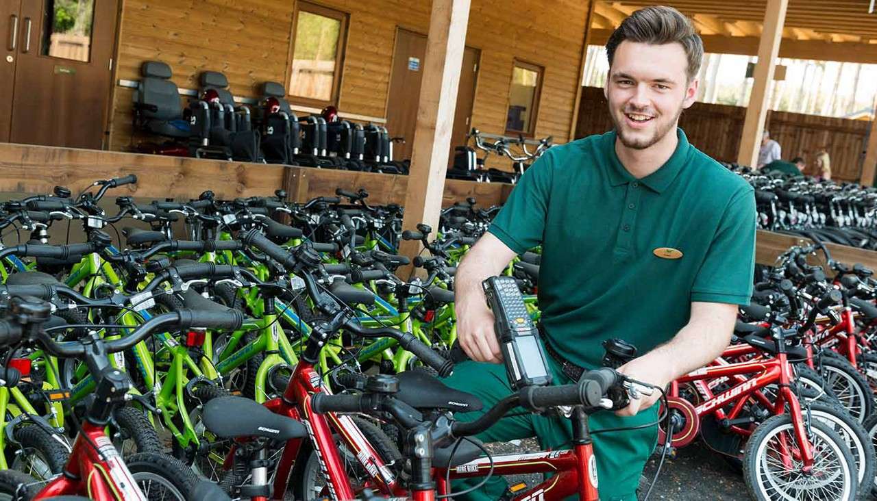 Attendant steadies a red child’s bicycle and operates a handheld scanner, smiling. Surrounding rows of red and green bikes fill a wooden, open-air rental shop with helmets on racks.