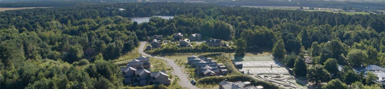 Cabin-style lodges nestle among trees, linked by winding roads; nearby tennis courts and a curving go-kart track lie open, with a lake and dense forest stretching across the countryside.