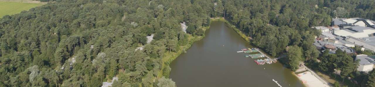 Lake sits calm, bordered by dense forest; small docks with paddleboats line the right shore near a sandy beach and buildings, while tree-covered hills surround the waterway.