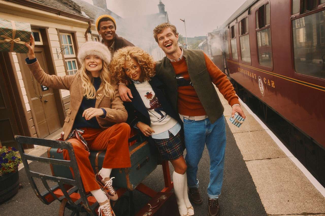 Four fashionably dressed travelers pose and laugh on a luggage cart; one holds a gift. They wait beside a maroon train car emitting steam. Visible text: "RESTAURANT CAR".
