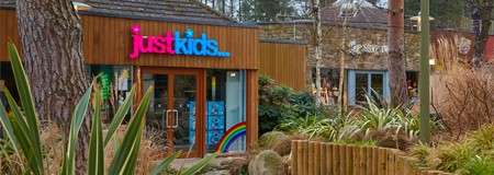 Shopfront displays sign reading "just kids..." above glass double doors; window shows colorful graphics including a rainbow; plants, trees, and wooden cladding surround the building in a landscaped outdoor setting.