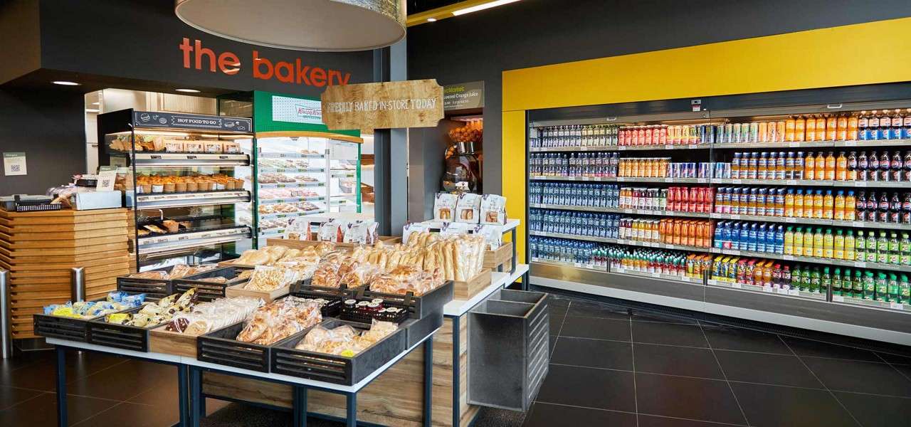 Bakery goods sit displayed on a central table, inviting purchase in a modern store with refrigerated drinks along the wall. Text: the bakery; HOT FOOD TO GO; FRESHLY BAKED IN-STORE TODAY.