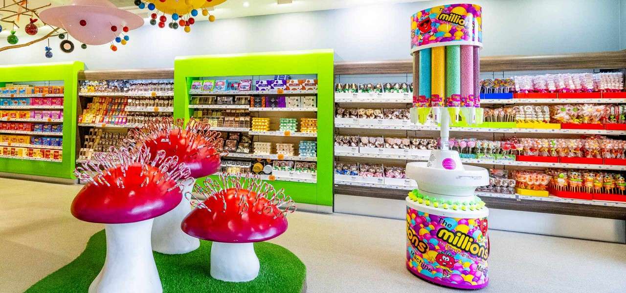 Candy dispenser displays multicolored pellets in a bright candy shop; nearby red mushroom stands hold lollipops; shelves of packaged sweets line the walls. Text: “The millions”, “Machine”.