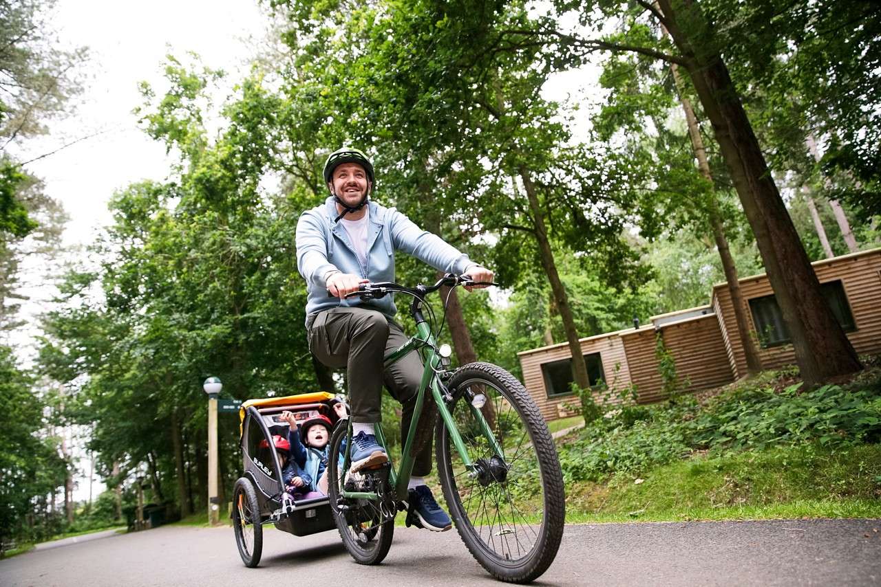 Adult cyclist pedals a green bike, towing a child trailer with two kids waving; context: paved forest path beside wooden cabins. Visible text: BURLEY.