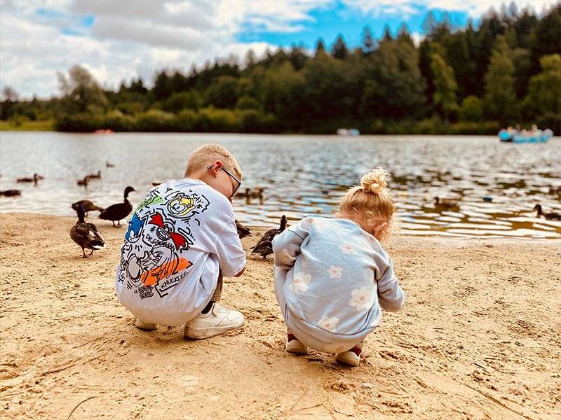 Two children crouch, playing in sand and watching ducks. A sandy shore meets a calm lake with swimming birds, pedal boats, and a forested tree line under partly cloudy skies.