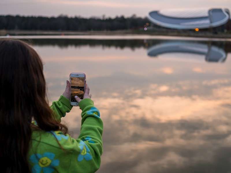 Person with long hair holds a smartphone, photographing calm lake at sunset; clouds reflect on water, distant modern curved structure across shoreline, soft light and trees framing quiet scene.