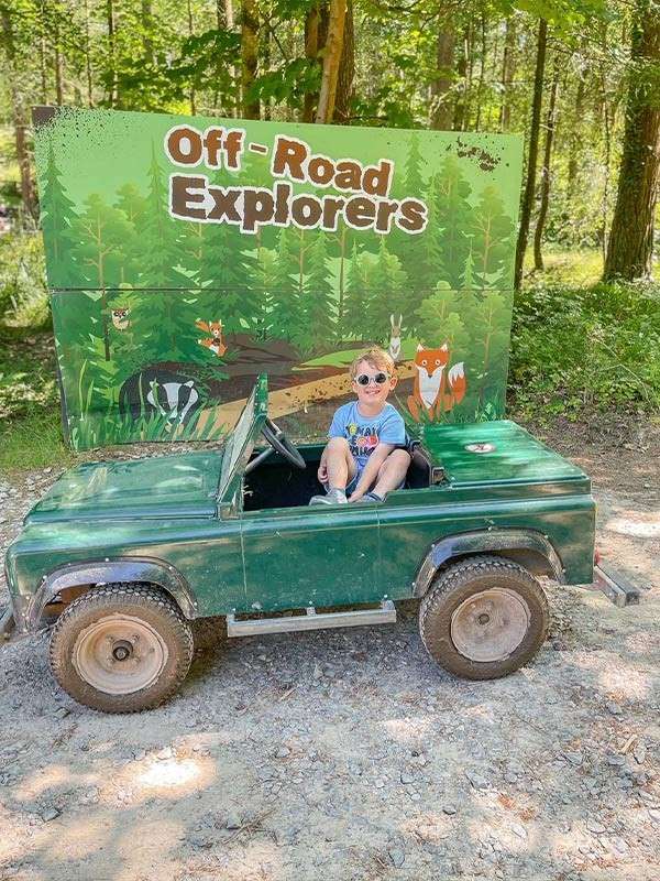 Child sits in a small green toy jeep, gripping the wheel; behind, a forest-themed board reads: Off-Road Explorers. Scene set on a gravel path in sunlit woods.