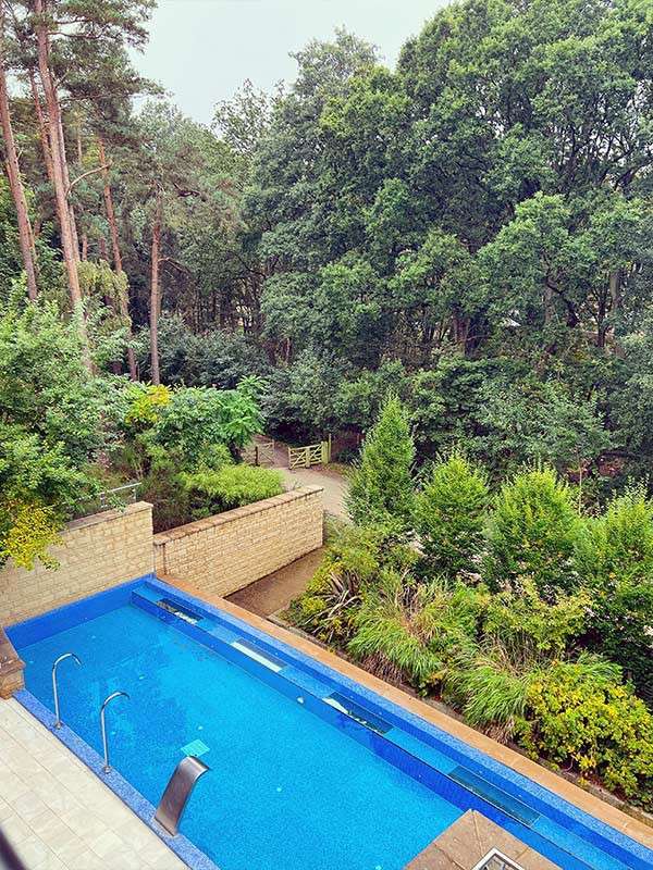 A narrow blue swimming pool stretches along a patio, with metal fountains and steps; beyond a low stone wall, a path and wooden gate lead into dense green woodland.