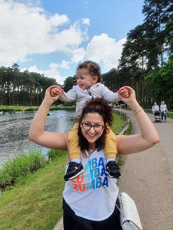 Woman carries a toddler on her shoulders, holding the child’s hands; both smiling. On a lakeside park path with trees, water, and people in the background. Shirt text: ZUMBA ZUMBA ZUMBA.