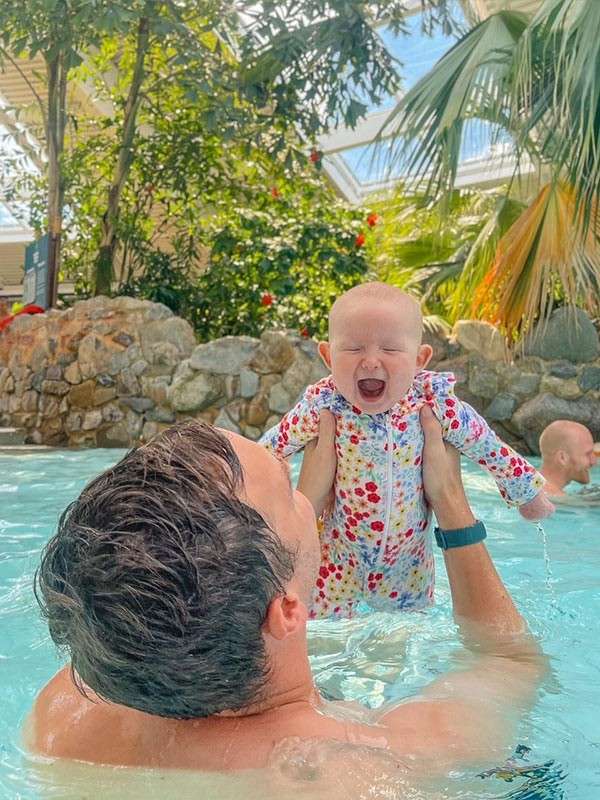 Baby in a floral swimsuit squeals with mouth open while being lifted from the water by an adult. Tropical indoor pool features palm trees, rock wall, skylight, and another swimmer.