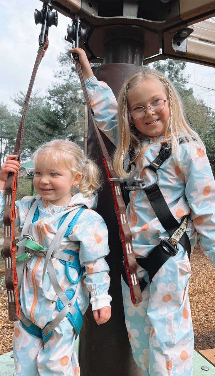 Two young girls smile and hold safety straps while secured in harnesses, standing under a pole at an outdoor play structure, wearing matching floral suits; trees and mulch surround them.