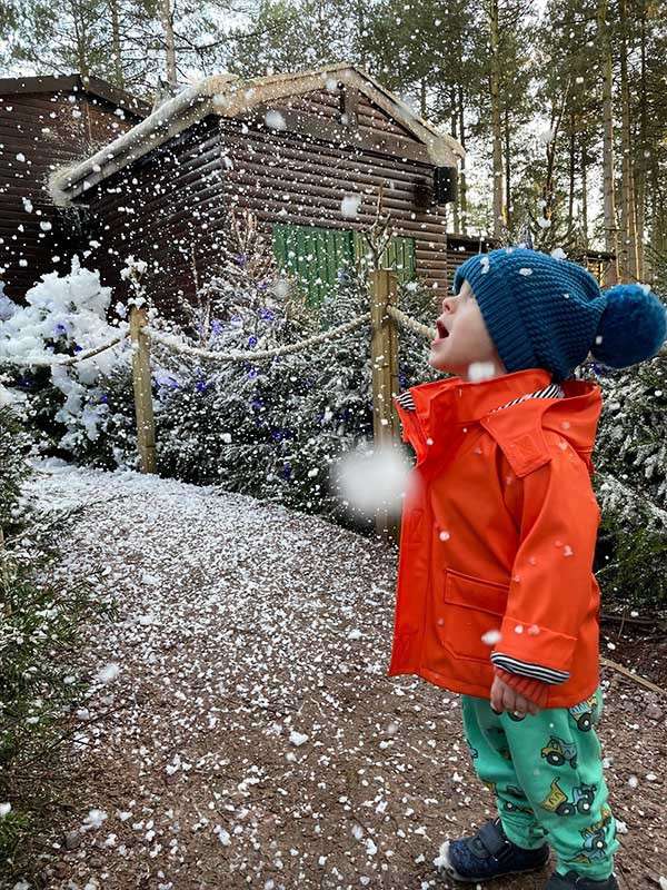 Child in orange raincoat and blue pom-pom hat gazes upward, mouth open, as snow falls. Surrounded by snowy firs, rope fence, and a log cabin along a forest path.