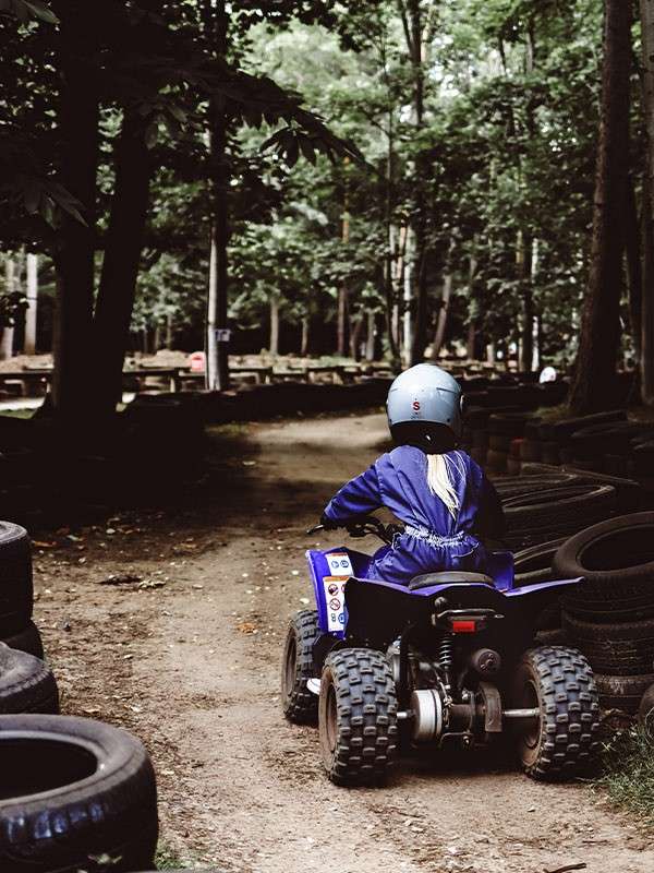 Child on a quad bike rides forward, wearing a helmet marked "S". The vehicle follows a dirt path through a forested track bordered by stacked tires and wooden barriers.