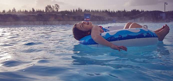 Child reclines on a blue inflatable tube, smiling and drifting. The tube floats in a calm outdoor pool, with misty water, rocky edging, distant trees, and a cloudy evening sky.