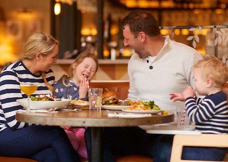 Two adults and two children laugh while eating at a round restaurant table; plates of salad, burgers, and drinks under warm lighting, with a toddler in a high chair.