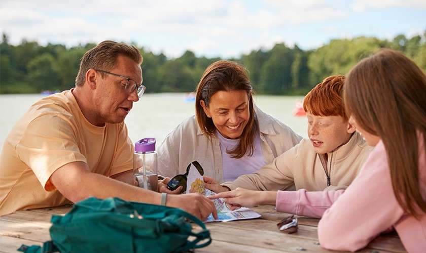 Family of four studies a map, pointing and discussing, at a wooden picnic table. Surroundings: lakeside park with trees, backpack, water bottle, binoculars, and calm water in the background.