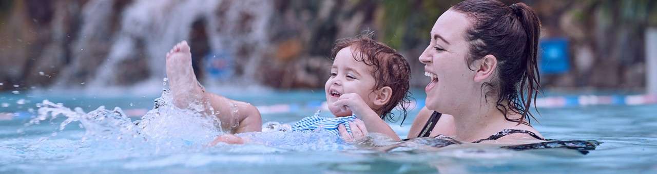 Adult and toddler splash and laugh; the child kicks water while being held. Context: a swimming pool with blurred waterfall rocks and a lane rope in the background.