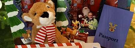 Plush reindeer toy in a red-and-white scarf sits amid a Christmas display: paper trees, candy, reindeer figurines, a photo of someone in a Santa costume, and a booklet labeled: Passport.