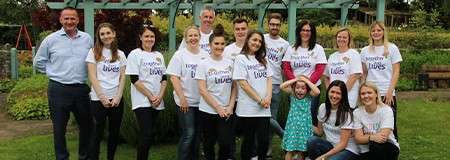 Group of volunteers pose smiling, wearing matching white T-shirts reading “Improving Lives,” standing in a garden under a pergola with greenery, benches, and a child in front.