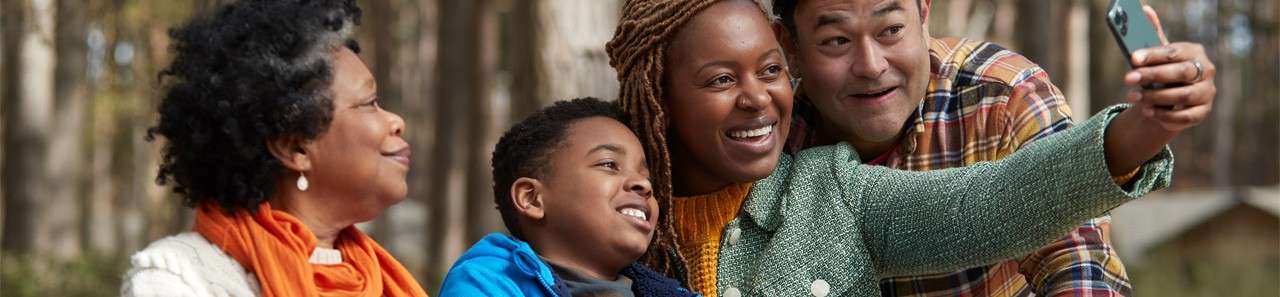 Four people pose, smiling, as one holds a smartphone to take a selfie, sitting closely together in a forested park with soft daylight filtering through trees.