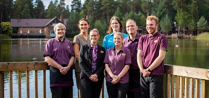 Eight uniformed workers stand posing and smiling for a group photo, hands clasped, on a wooden lakeside deck, with forest trees and cabin buildings across the water in daylight.