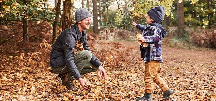 Child tosses fallen leaves toward a crouching adult, both wearing warm hats, on a leaf-covered woodland path surrounded by trees in autumn.
