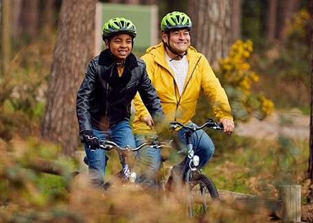 Two cyclists ride forward, wearing green helmets; one adult in a yellow jacket, one child in a black jacket, both smiling; context: a forested trail with trees, shrubs, and soft sunlight.