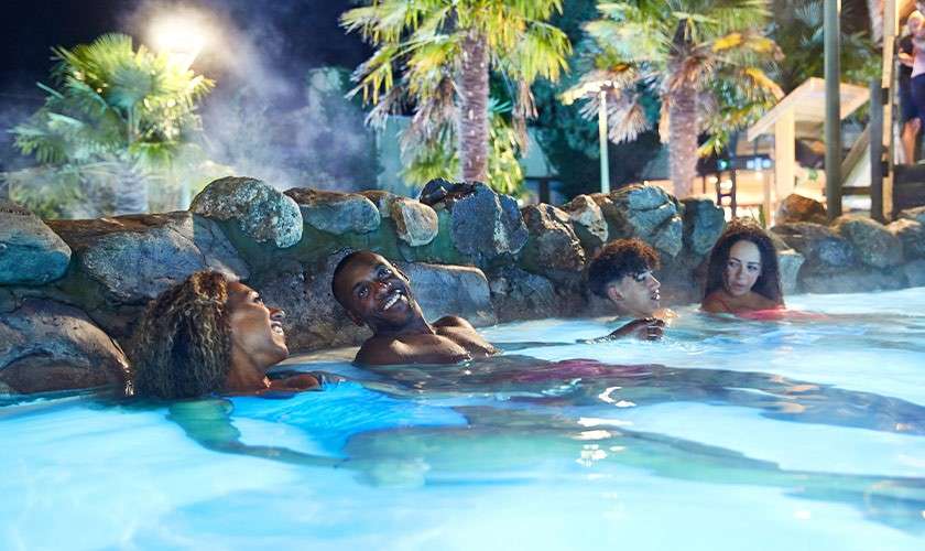 Four people relax and chat, half-submerged in a steaming outdoor pool at night, leaning against a rock wall; palm trees and soft lights surround the tropical-style spa setting.