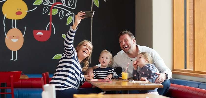 Family poses for a selfie; mother holds a smartphone while father and two young children smile. Context: bright diner with red booths, wooden tables, wall art, large window, and orange juice.