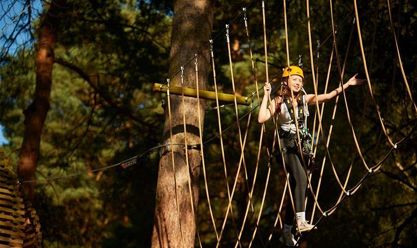 Participant wearing helmet and harness crosses a rope bridge, stepping carefully and holding support lines, in a treetop adventure course among tall pine trees, with sunlight filtering through the forest.