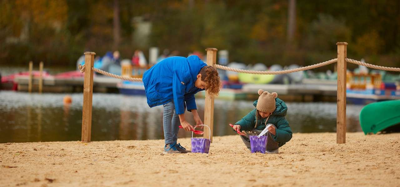 Two children dig with red spades, filling purple buckets. They crouch on a sandy beach beside a lake, bordered by rope posts, with paddleboats and autumn trees blurred in background.