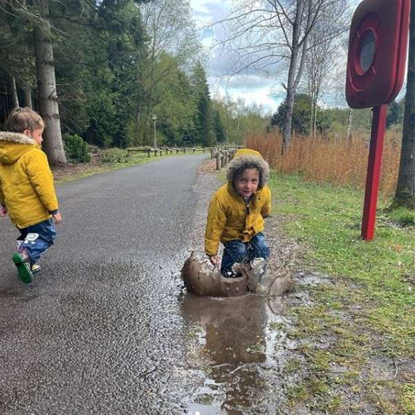 Child in yellow coat splashes in a muddy puddle on a paved park path, water spraying; another child walks nearby. Tall trees, grass, and a red post frame the scene.