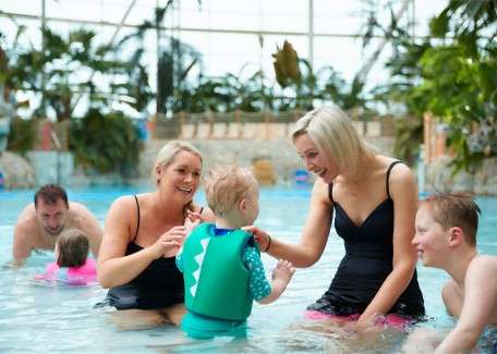 Child in green flotation vest splashes as two adults guide and smile; another child watches. Other swimmers play in a shallow indoor pool bordered by plants beneath a glass roof.