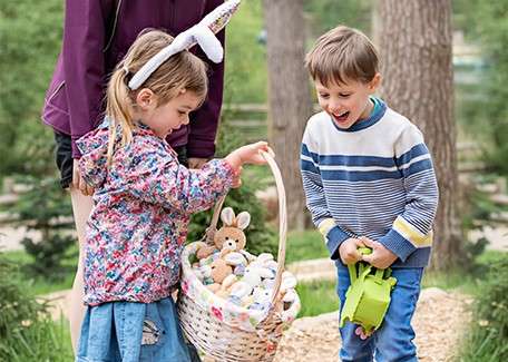 Two children smile as the girl lifts a white basket filled with plush bunnies, while the boy watches holding a green watering can, outdoors on a wood-chip path among trees.