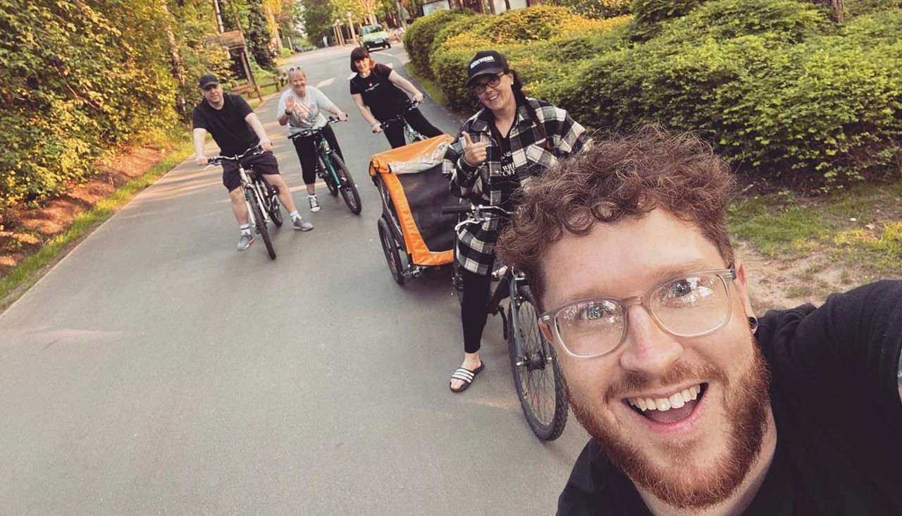 Cyclists ride together; front rider takes a smiling selfie while others follow, one towing an orange child trailer; scene unfolds on a quiet, tree-lined residential street in warm daylight.
