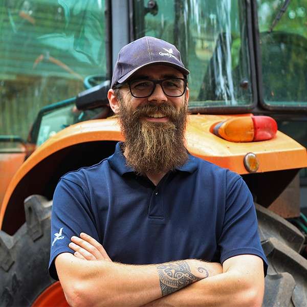Bearded person in glasses smiles with arms crossed, standing before an orange tractor. Outdoor setting with trees reflected in the cab windows; navy cap and polo, forearm tattoo visible.