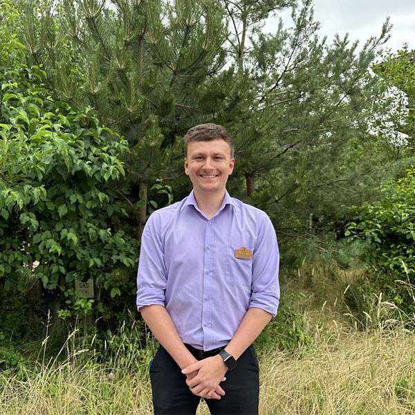 Smiling man stands with hands clasped, wearing a lavender shirt and black pants, in tall grass before pine trees under overcast sky. Visible sign reads: “GAS”.