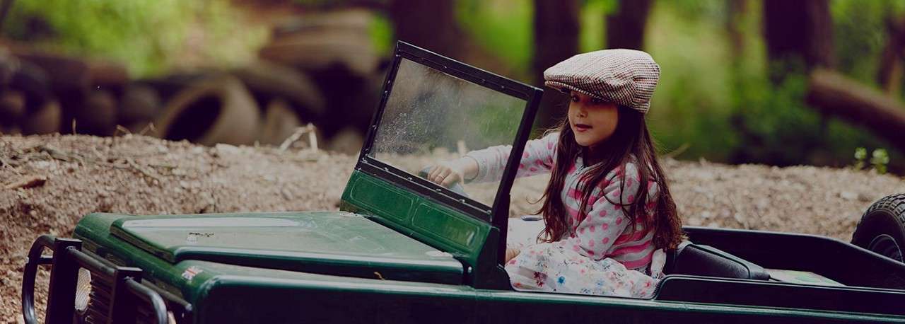 Child wearing a flat cap sits in a small green toy jeep, hands near the wheel, in a wooded area with dirt ground and stacked tires in the background.
