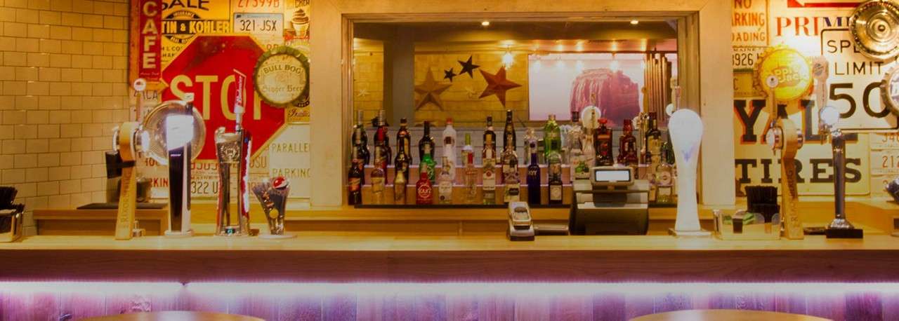 Bar counter displays beer taps and liquor bottles in a retro pub, surrounded by tiled walls and vintage signs. Visible text: CAFE; STOP; NO PARKING; SPEED LIMIT 50; TIRES; 321 JSX.