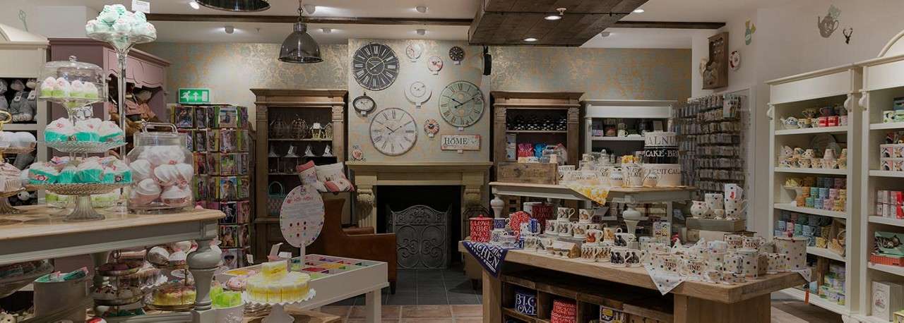 Gift-shop displays present mugs, teacups, and confections, arranged on wooden tables; context: cozy boutique interior with shelves, a fireplace, and wall clocks. Text: EXIT; HOME; BIG LOVE; LOVE; MINE.