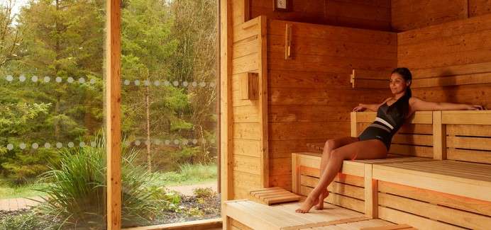 Person reclines on wooden sauna bench, relaxing with arms stretched. Sunlight filters through a large glass wall, revealing green trees and plants outside, creating a warm, tranquil spa setting.