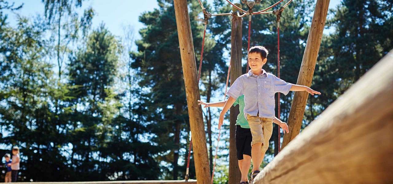 Two children balance-walk along a wooden beam, arms outstretched, on a ropes-and-poles playground, while other kids play in the background amid tall trees on a sunny day.